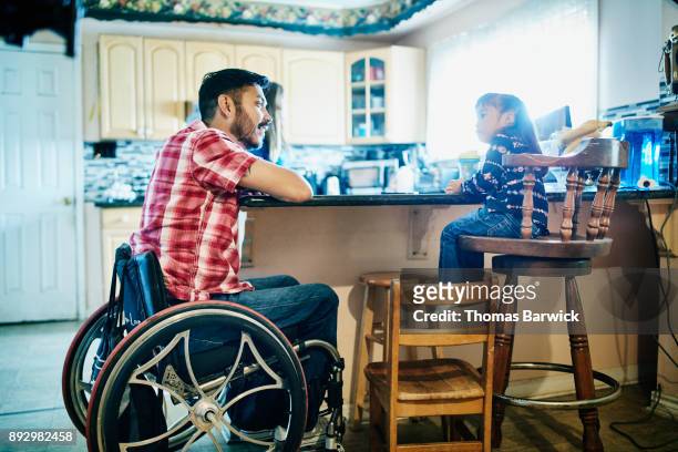Smiling father using a wheelchair in discussion with young daughter while sitting together at kitchen counter.