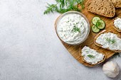 Homemade greek tzatziki sauce in a glass bowl with ingredients and sliced bread on a wooden board and light stone background. Cucumber, lemon, dill. Top view, horizontal image, copy space