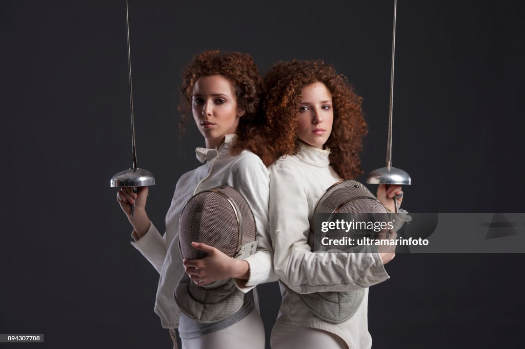 Fencing sport competition. Duel. Dark grey background. Sporting Women. Two curly redhead twin girls fencer. Twins.