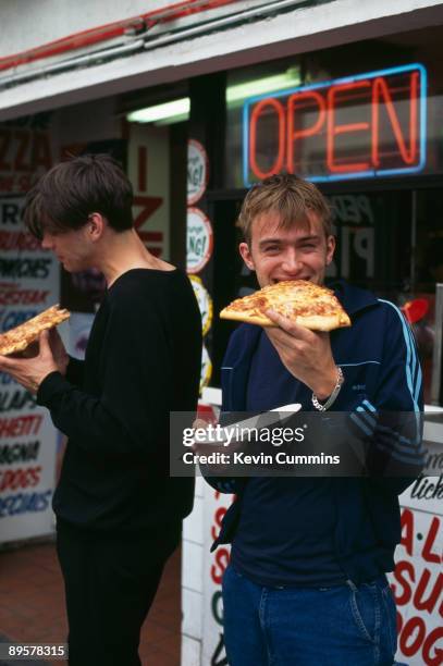 Bassist Alex James and singer singer Damon Albarn, of English pop group Blur, eating pizza outside a snack bar, circa 1995.