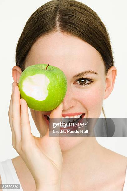 young woman holding green apple over eye, smiling - pomme croquée photos et images de collection