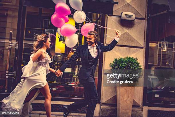 affectueuse mariée et le marié, main dans la main, heureusement courir dans la rue - wedding couple laughing photos et images de collection