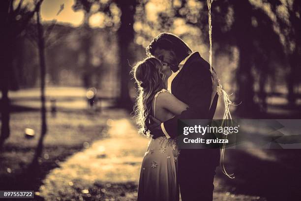 affectionate bride and groom, embraces and kisses amidst the gravel path at the park at sunset. - white dress stock pictures, royalty-free photos & images