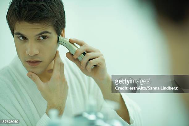 young man looking in mirror, shaving with electric razor - electric razor stock pictures, royalty-free photos & images