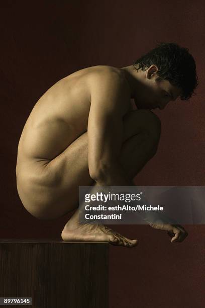 nude man crouching with chin resting on knees, side view - clair obscur stockfoto's en -beelden