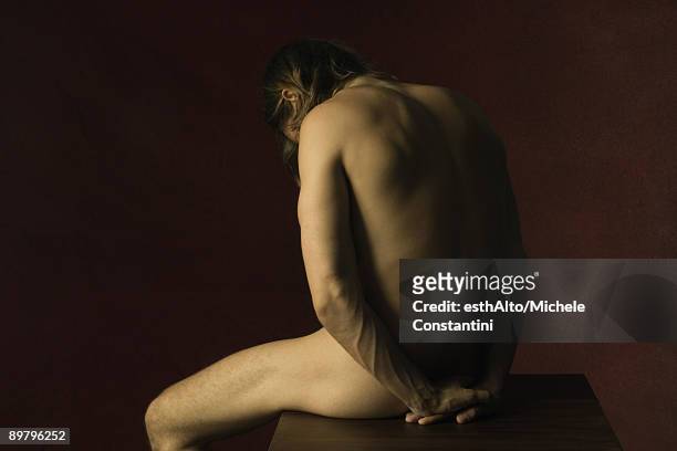 nude man sitting with hands clasped behind back, head down - clair obscur stockfoto's en -beelden