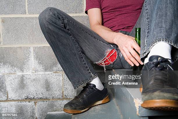 a man sitting on a step and holding a bottle of beer - ripped jeans stockfoto's en -beelden