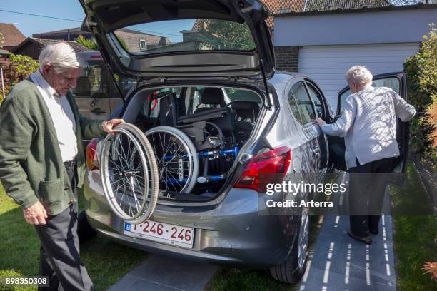 Senior husband fitting dismantled wheelchair from physically disabled elderly wife in car trunk at home before going for a ride.