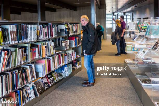 Elderly man choosing book to read from rows of books in bookshelves at public library.