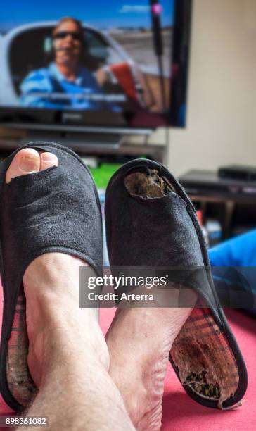 Couch potato, lazy man in comfy chair wearing worn slippers with big toes sticking through and watching television set in living room.