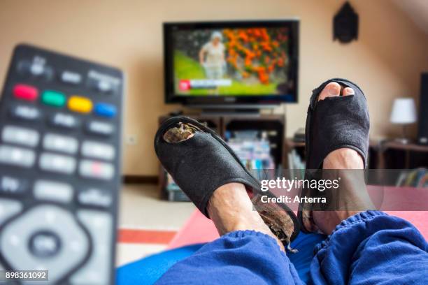 Remote control and couch potato, lazy man in comfy chair wearing worn slippers with big toes sticking through and watching television in living room.