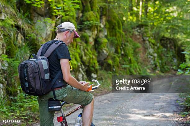 Cyclist with backpack riding a bicycle stopping to take a break for texting and reading smartphone along a forest road in summer.