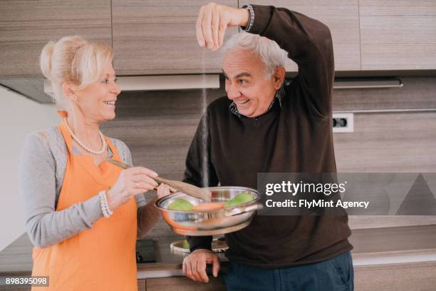 casal sênior na cozinha a fazer o jantar, o homem é adicionar o sal - adicionar sal - fotografias e filmes do acervo