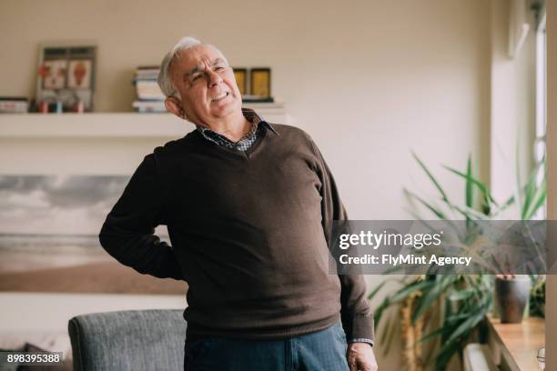 senior man in a daily room having a pain in his back - bending over backwards stock pictures, royalty-free photos & images