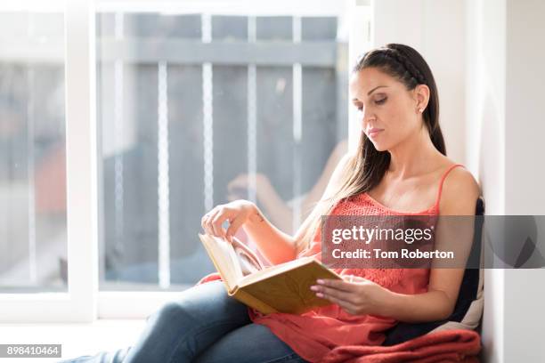woman sitting in window seat at home relaxing and reading book - tom roberton stock pictures, royalty-free photos & images