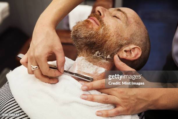 a customer sitting in the barbers chair, having his chin shaved by a barber using a cut throat razor. - barbier stockfoto's en -beelden