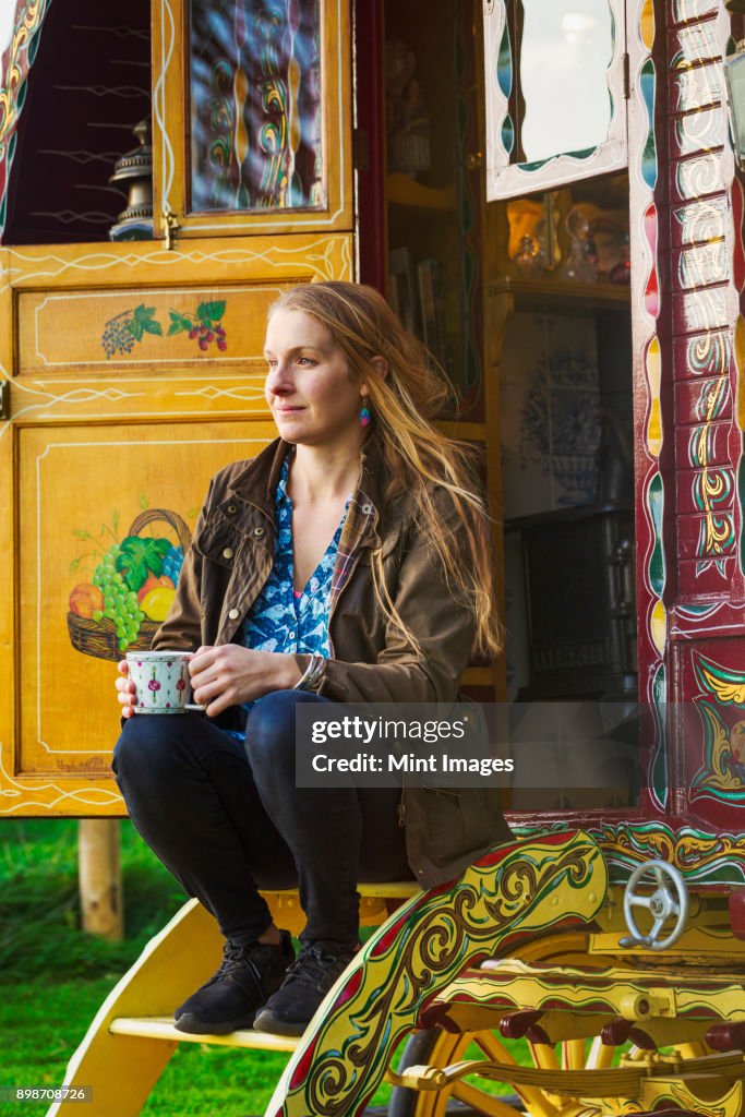 A woman seated on the steps of a caravan, holding a mug of tea.