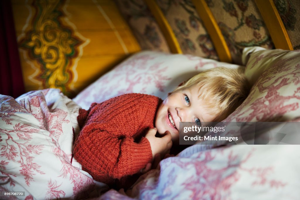 A boy lying in bed under a curved bow top roof of a gypsy caravan, looking at the camera.