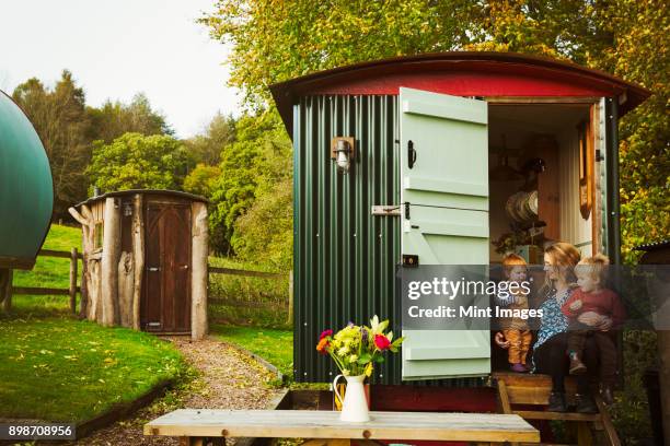 a shepherds hut with open door beside a path to a small rustic shed, and a woman with two small children seated on the step. - shed fotografías e imágenes de stock