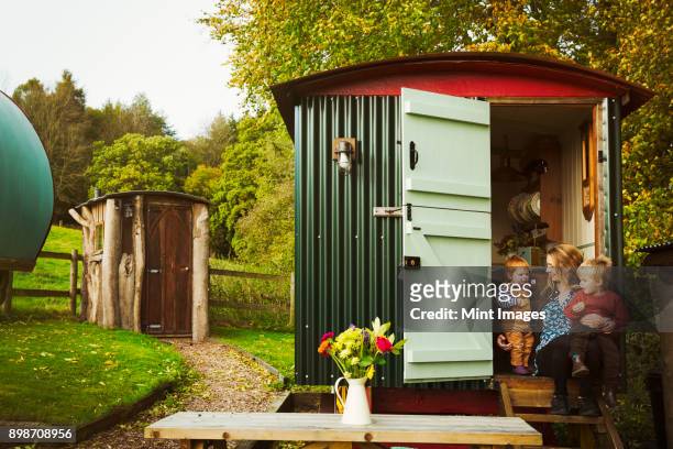 a shepherds hut with open door beside a path to a small rustic shed, and a woman with two small children seated on the step. - shed stock pictures, royalty-free photos & images
