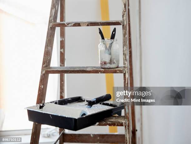 a wooden paint splashed ladder, a jar with two paintbrushes and a paint tray and roller. - verfbak stockfoto's en -beelden