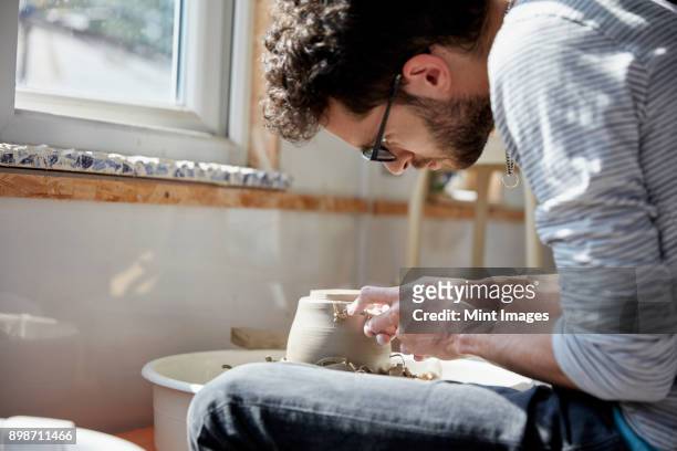 man seated at a potters wheel, moulding the wet clay on the spinning turntable, in a pottery studio - man pottery stock pictures, royalty-free photos & images