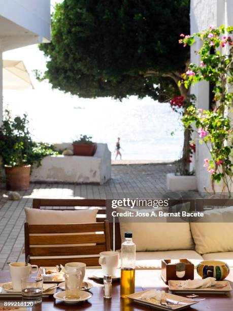 table of a terrace of bar in the street close to the sea, with the plates and glasses of a breakfast. to the bottom a person walks close to the beach. san jose, cabo de gata - nijar natural park, beach, almeria, andalusia, spain - almeria restaurant stock-fotos und bilder