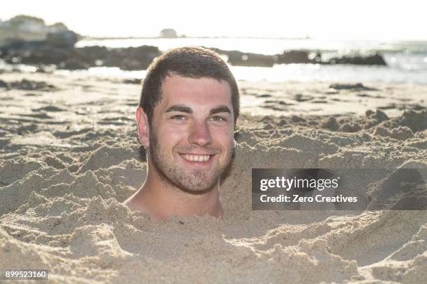 portrait of young man buried up to his neck in sand on beach - head in sand stock-fotos und bilder