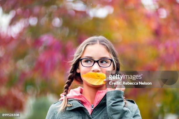 portrait of girl with plaits and glasses covering mouth with leaf looking away - leesbril stockfoto's en -beelden