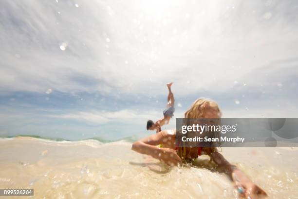 girl lying on beach in shallow water, brother doing handstand in background - destin beach stock-fotos und bilder