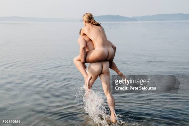 nude man giving nude woman piggyback into water - women skinny dipping stockfoto's en -beelden