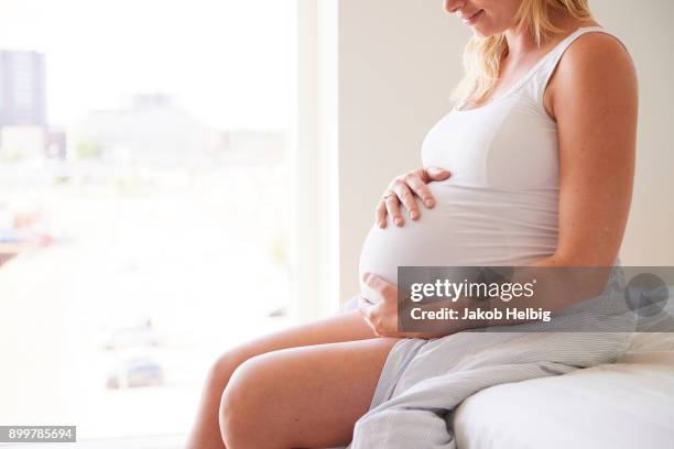 cropped shot of pregnant young woman sitting on bed with hands on stomach - jakob helbig stock pictures, royalty-free photos & images