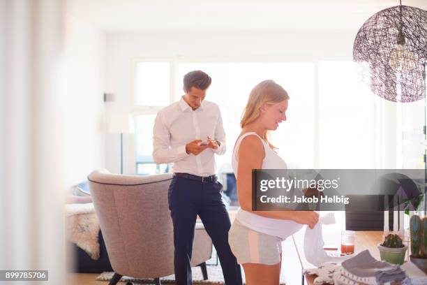 man looking at smartphone and pregnant girlfriend folding laundry in living room - jakob helbig stock pictures, royalty-free photos & images