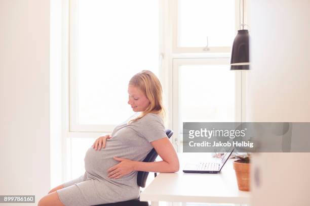 pregnant young woman at desk gazing at stomach - jakob helbig stock pictures, royalty-free photos & images