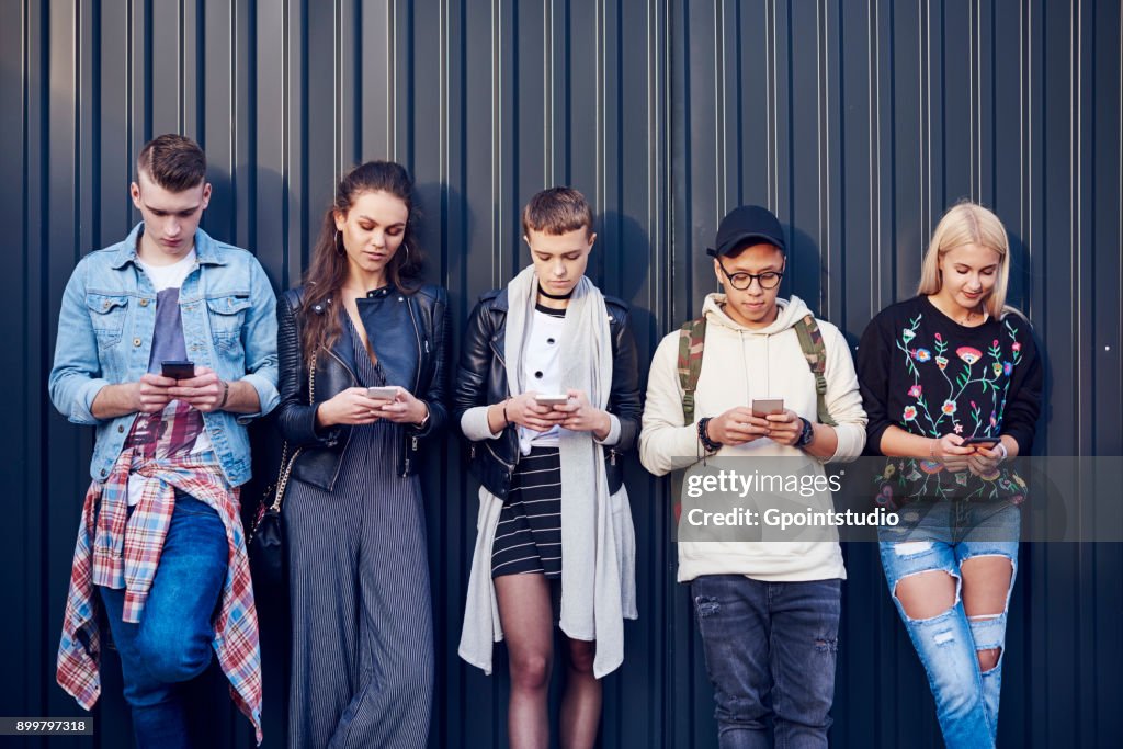 Row of five young adult friends leaning against black wall looking at smartphones