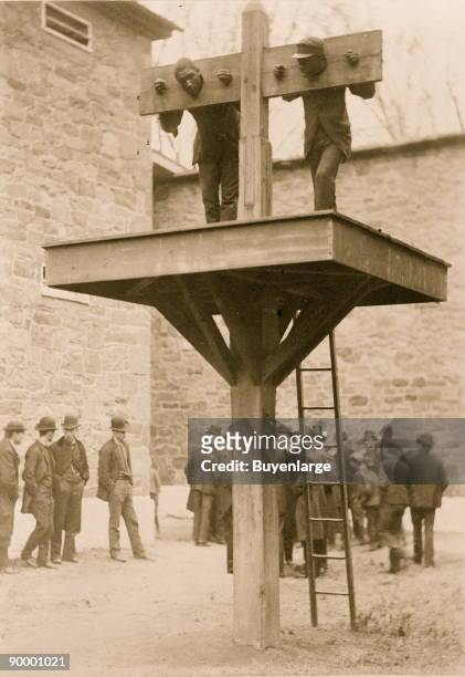 Ground level whipping post, above which is a platform for the pillory, presently occupied by two African American men; a ladder leans against the...