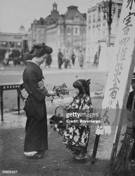 Toddler in front of Kanban Shop sign sells flowers to a kimono wearing young lady who picks them from a basket that the little girls is holding.
