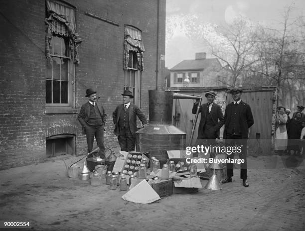 Onlookers watch as suited men stand in front of a large copper kettle still for making illegal liquor with boxes of bottles and funnels spread before...