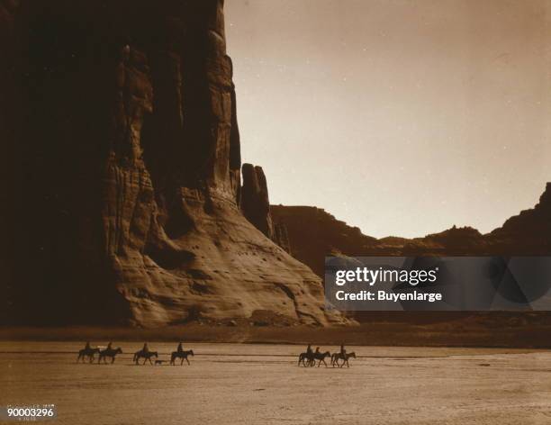 Seven riders on horseback and dog trek against background of canyon cliffs.