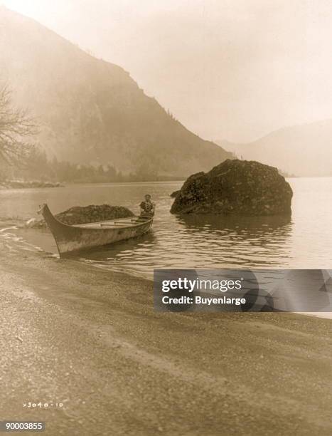 Chinook man seated in rear of boat beached on riverbank, mountains in background.