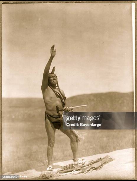Dakota man, wearing breechcloth, holding pipe, with right hand raised skyward.