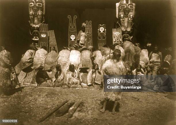 Kwakiutl people, some bowing before totem poles in background, others seated facing front as part of the nunhlim ceremony, the four days prior to the...