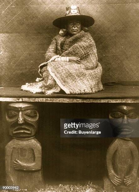 Kwakiutl woman, full-length portrait, seated on a blanket-covered board supported by two wooden carved images representing her slaves, facing front,...