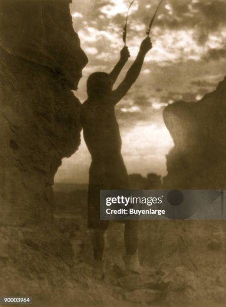 Bare-chested Tewa man with hands raised skyward, each holding a feather, as he faces the sun.