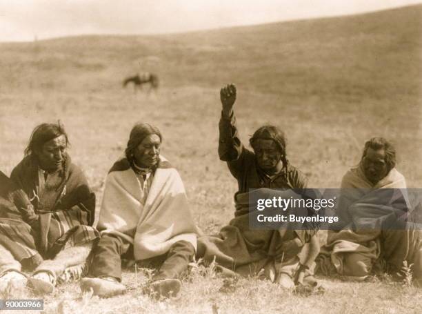 Four Atsina men, each wrapped in a blanket, seated on ground, one with arm and fist raised skyward, one holding a wing.