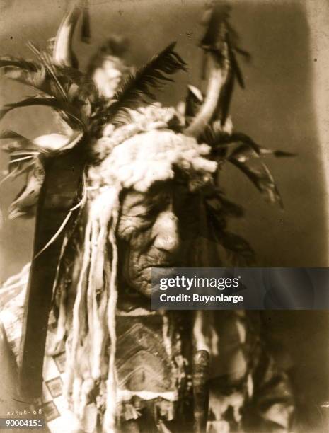 Lean Wolf, head-and-shoulders portrait, facing right, wearing horn headdress trimmed in white fur and dark feathers, beaded buckskin shirt.