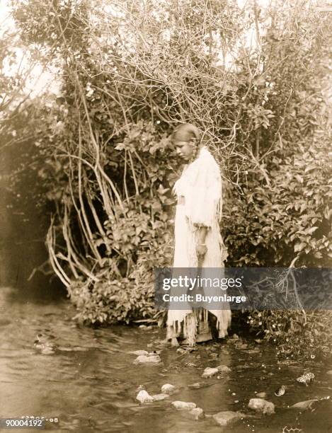 Young Arikara Indian standing in shallow water, wearing buckskin dress, with trees in background, North Dakota.