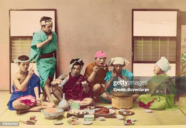 Men relax while eating and being serenaded by one member playing a stringed instruent like a guitar.