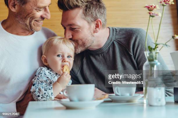 gay couple cuddling with their baby in cafe - gay stock-fotos und bilder