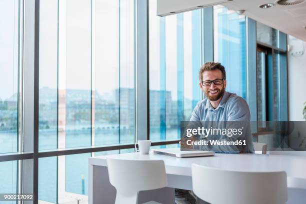 portrait of smiling man with laptop and cup of coffee sitting in modern office - barhocker stock-fotos und bilder